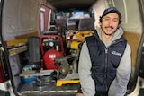 Carpenter Luke Kyriakides sits in the back of a white van wearing a puffer vest and cap, with tools behind him.