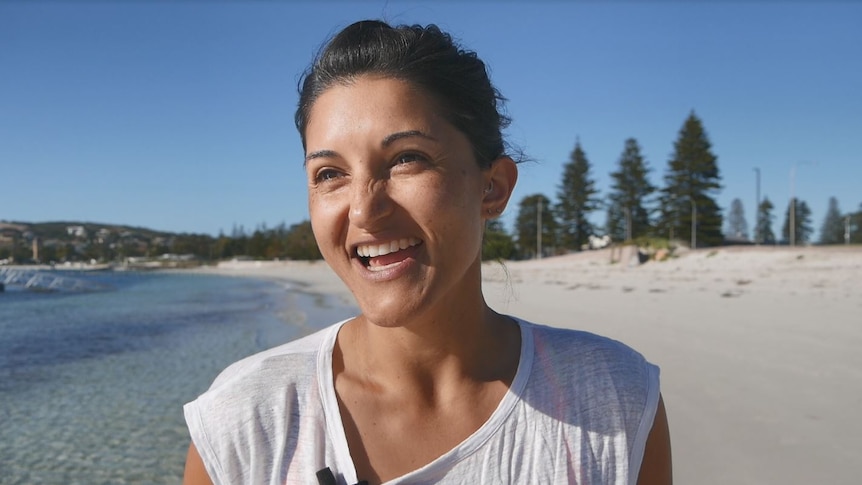 A close up, she wears a white t-shirt and has hair tied back. One side is the ocean, the other side is beach
