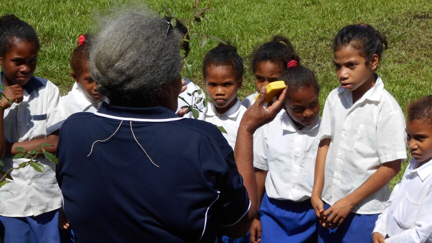 A lady with grey hair faces a group of children standing outside on grass  holding up a bar of soap