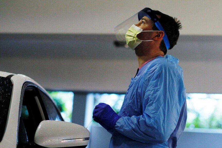 A man in protective face mask and scrubs stands next to a car