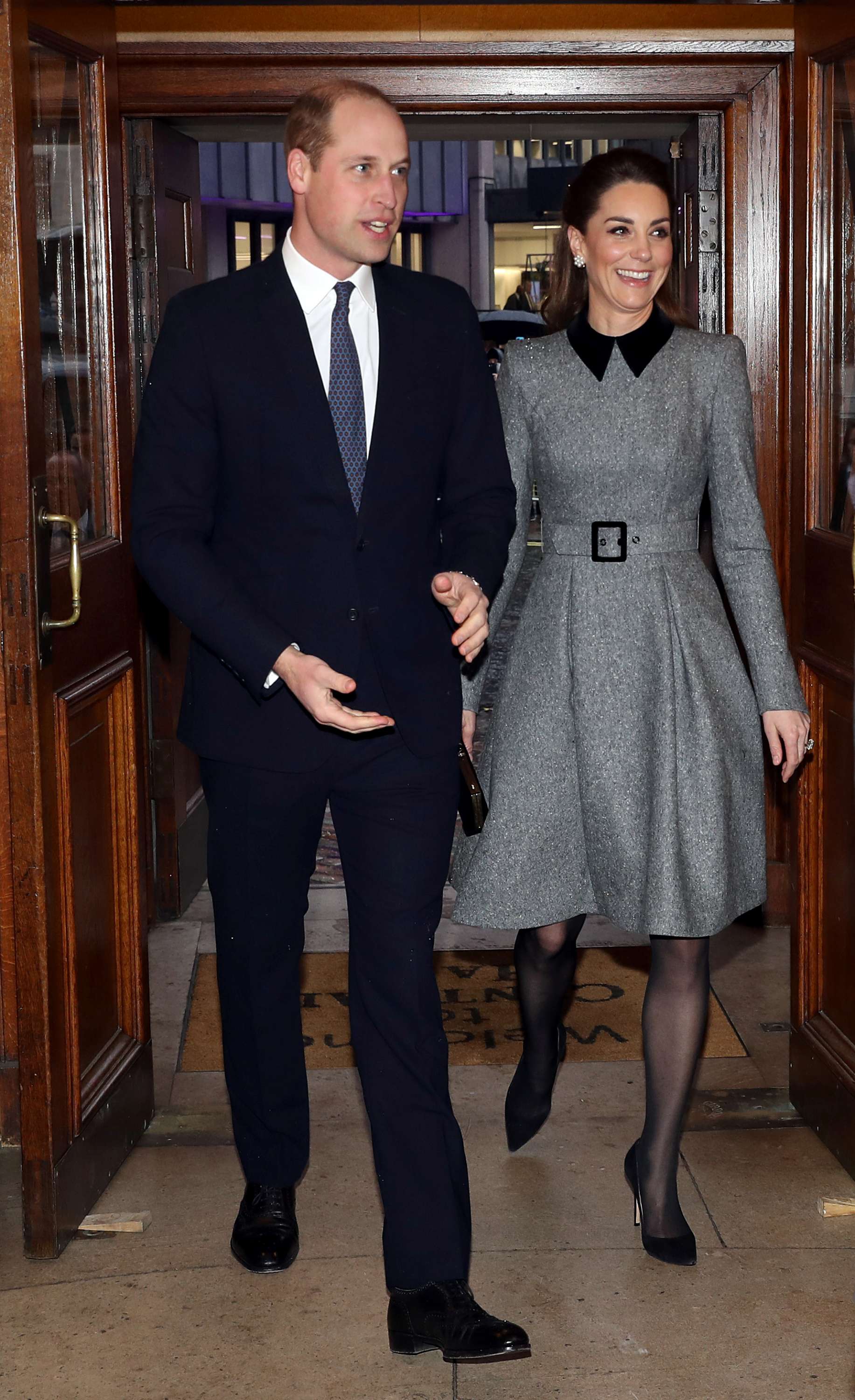 A man in a suit and tie and a woman in a grey dress hold hands as they walk through a hallway.