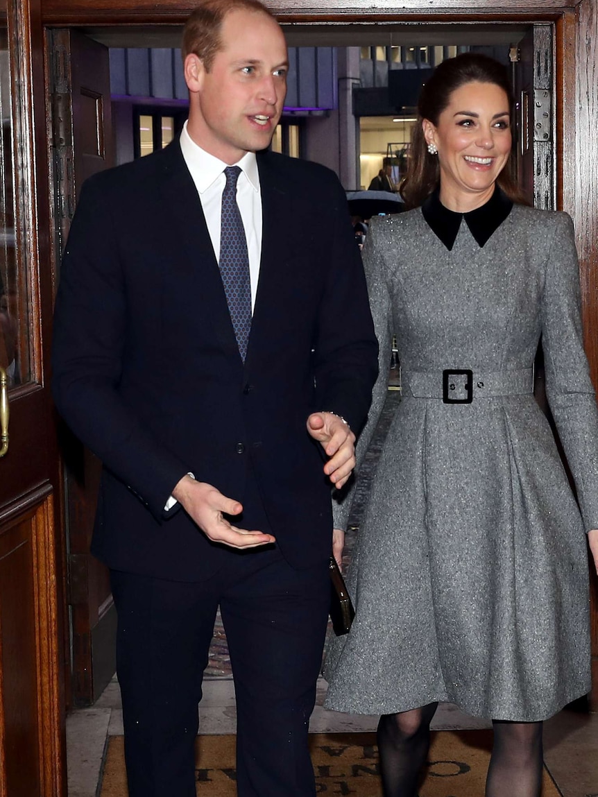 A man in a suit and tie and a woman in a grey dress hold hands as they walk through a hallway.