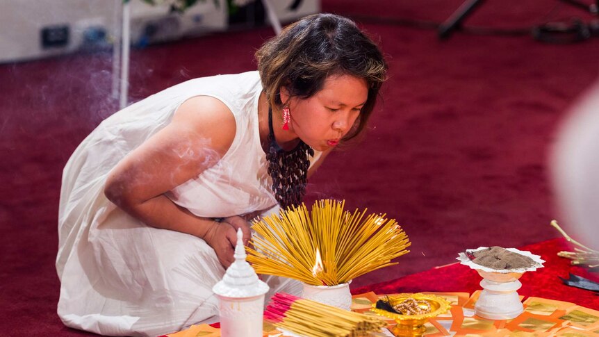 An asian woman kneels on red carpet and blows on a stack on incense sticks