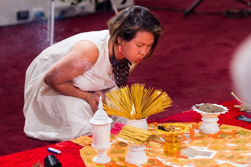 An asian woman kneels on red carpet and blows on a stack on incense sticks