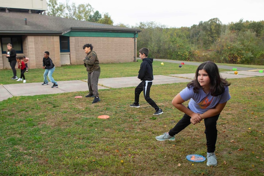 Seven primary students stand-spaced apart in a row, and they stretch to a side during an outdoor physical education class.