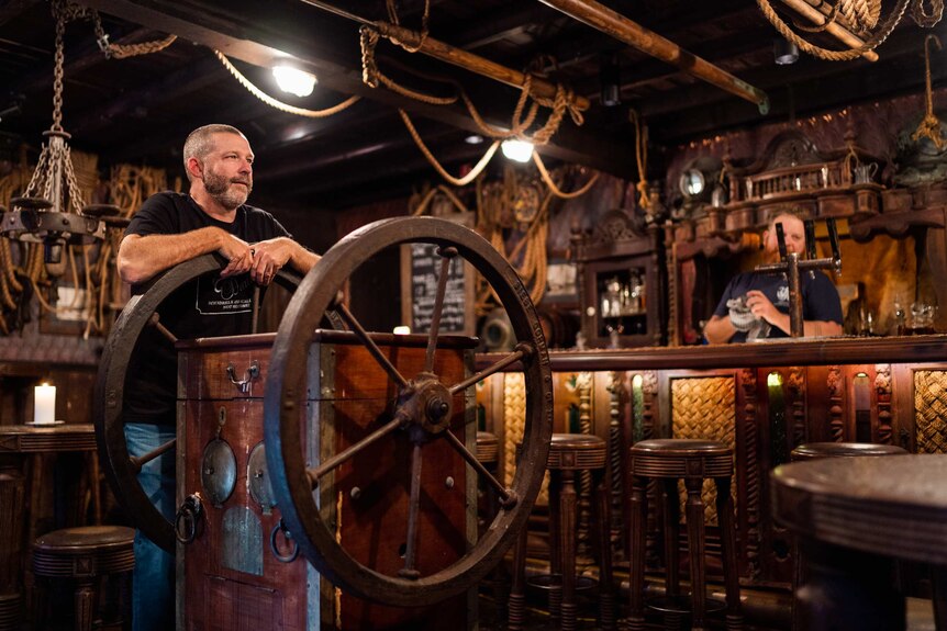 A man leans up against a ship's wheel in a pub with a bartender behind the bar nearby.