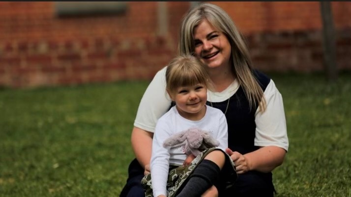 A mother sits on the grass with her infant daughter in her lap as they both smile at the camera.