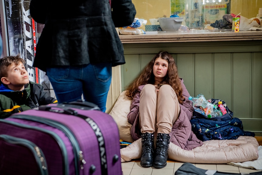 A young teen girl in black boots and a purple coat sits on the floor of a train station with a boy and a woman standing above 