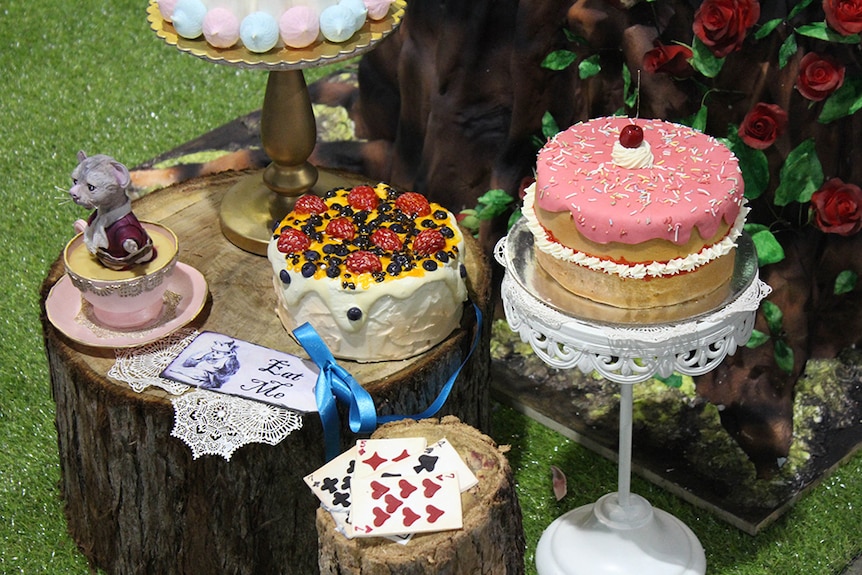 Sugar sculptures of Alice in Wonderland's Dormouse in a teacup beside three decorates cakes and playing cards.