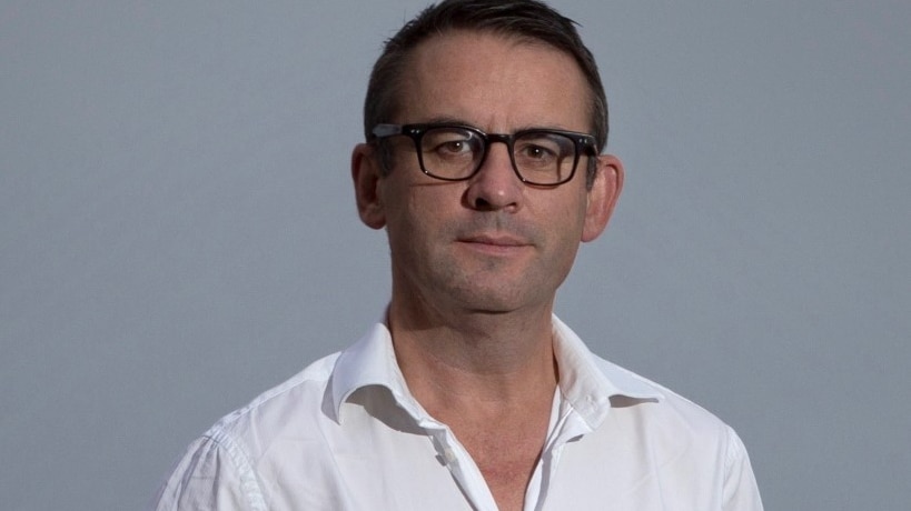 Paul Kildea standing in front of a grey background wearing black glasses and a white shirt.