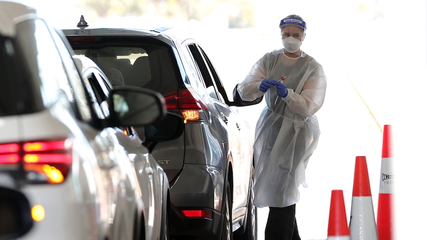 A testing site worker in full PPE takes a swab from a person at a drive-through testing site.
