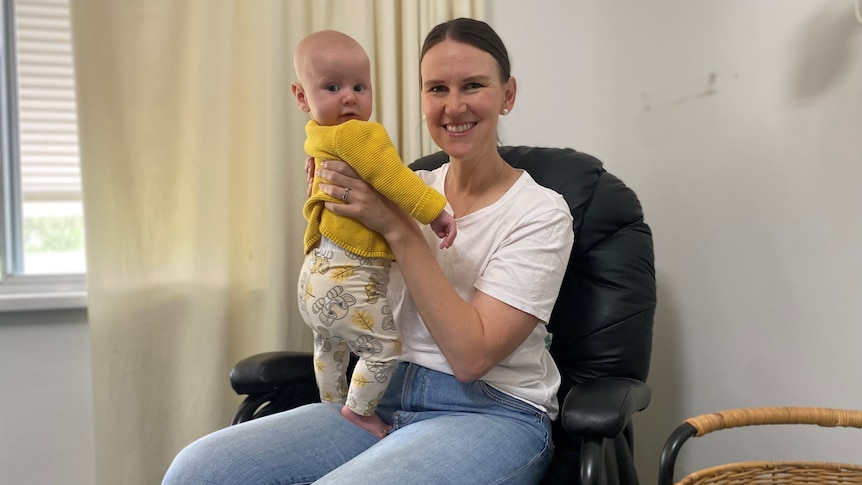 a woman in a white shirt and blue jeans holds her baby on her lap, both are looking at the camera