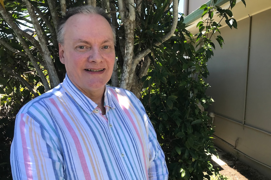 An older man, smiling, balding, wears pink, blue, green,yellow striped shirt, stands in front of tree.