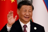 Xi Jinping stands in front of a Chinese flag smiling and waving.
