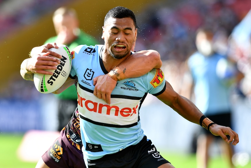 A Cronulla Sharks NRL player holds the ball with his right hand as he is tackled from behind by a Brisbane Broncos opponent.