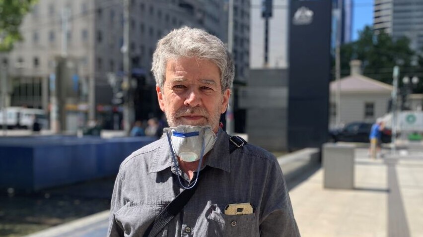 A man with grey hair and a face mask around his neck stands on a city street.
