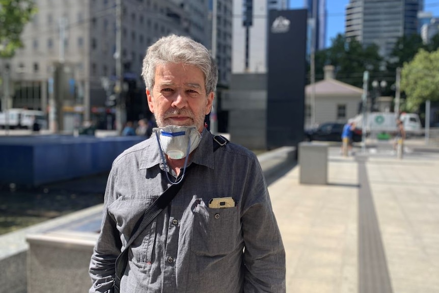 A man with grey hair and a face mask around his neck stands on a city street.