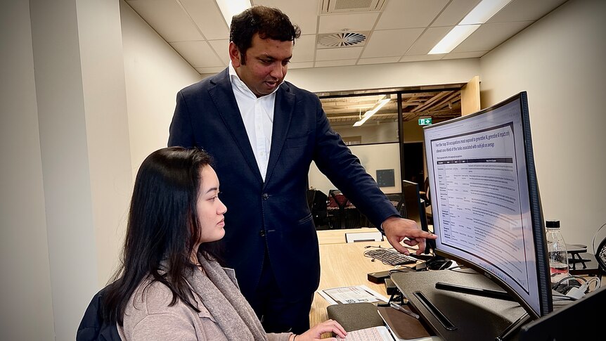 A man of colour in a blue suit and white shirt stands and points at a computer screen where a young Asian woman is working.