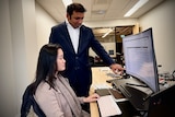 A man of colour in a blue suit and white shirt stands and points at a computer screen where a young Asian woman is working.