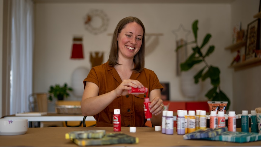 A young woman pours one bottle of red acrylic paint into another. Several other tubes of paint are scattered on the bench