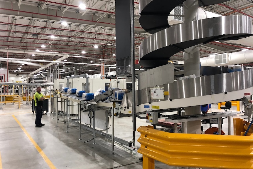 A man stands in the background watching packets of toilet paper move along a conveyor in a factory