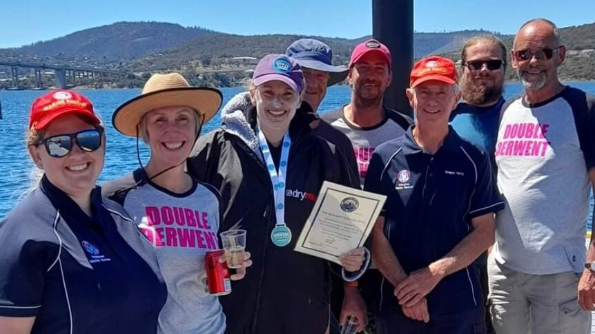 A group of people wearing caps including a woman with a medal and certificate in front of a river and hills.
