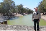A man wearing a khaki shirt and hat stands in front of the Darling River.