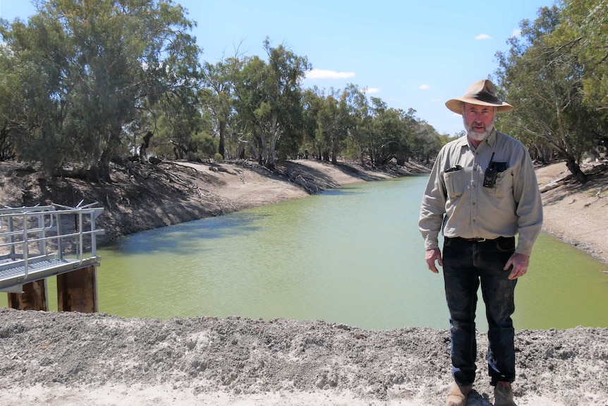 A man wearing a khaki shirt and hat stands in front of the Darling River.