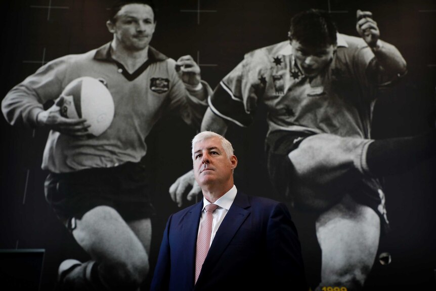 A man in a suit poses in front of a large wall photo of two rugby union players.