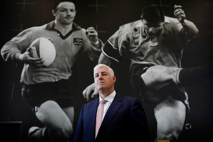 A man in a suit poses in front of a large wall photo of two rugby union players.