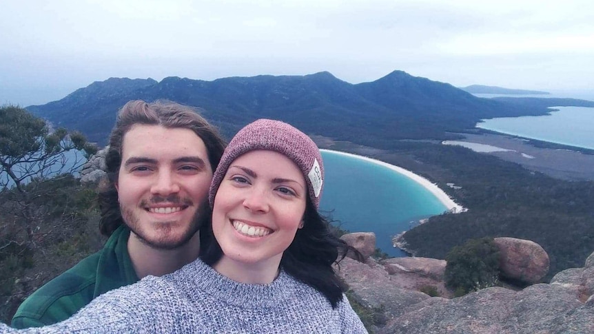 A smiling young couple dressed warmly, taking a selfie on Mt Amos, with Wineglass Bay showing in the background.