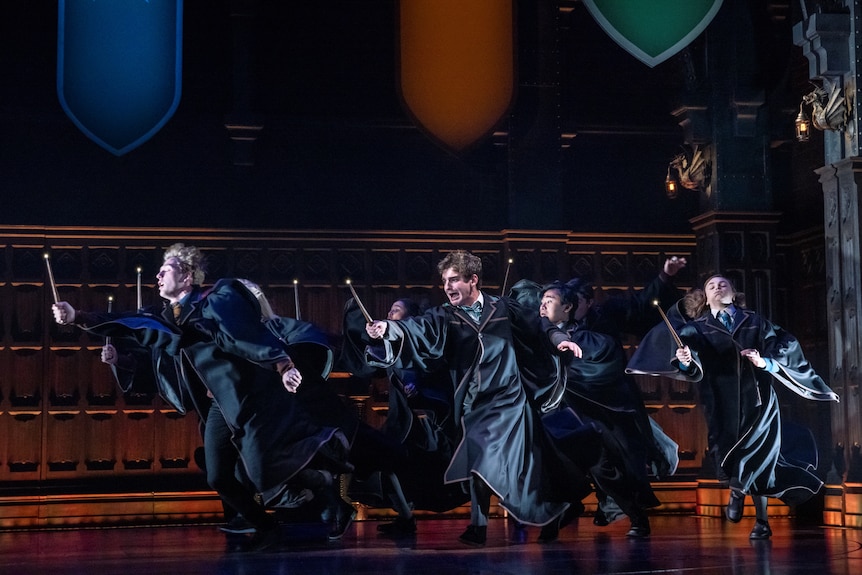 A group of people in dark robes run across a stage, all brandishing magic wands