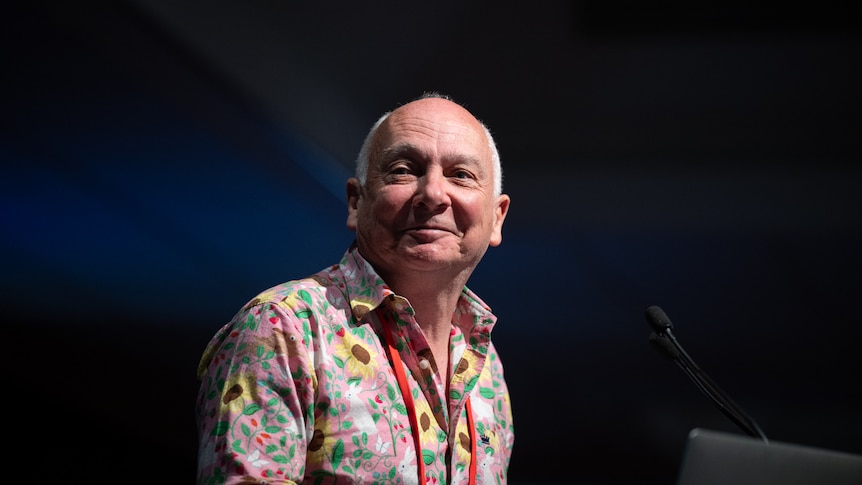 A man in front of a lecturn with white receeding hair and a pink sunflower shirt
