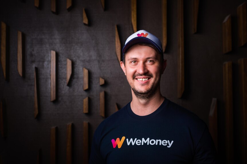 Man wearing a navy shirt with a logo for 'WeMoney' and a cap, standing in front of a dark wall.