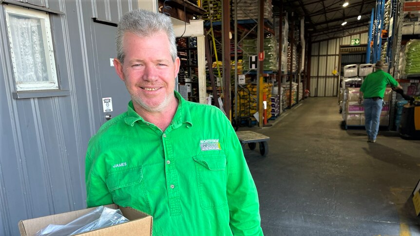 Mid shot of a man in a green shirt holding a box with a warehouse behind him 