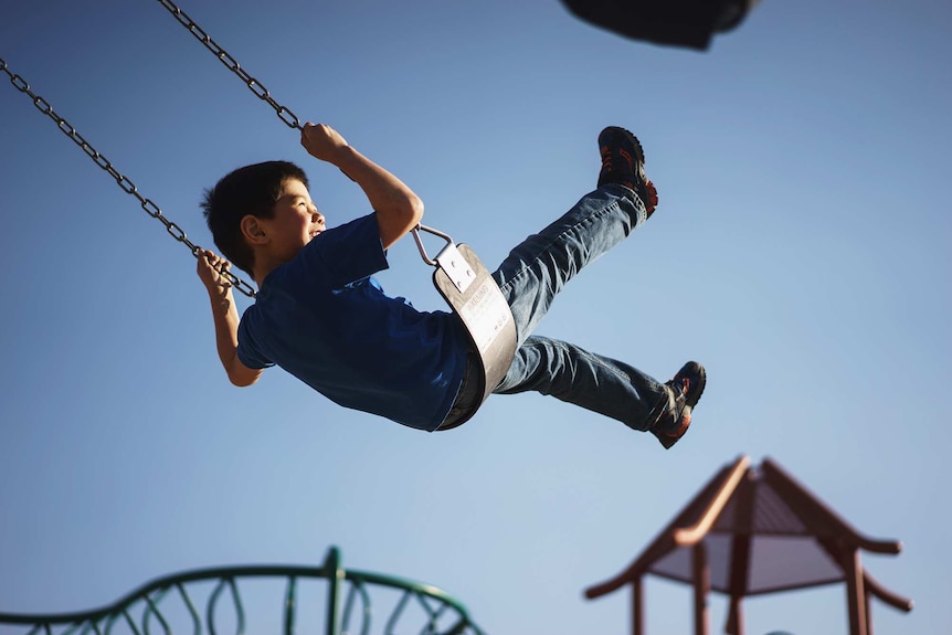 Young boy grinning while swinging high on a swing in a playground.