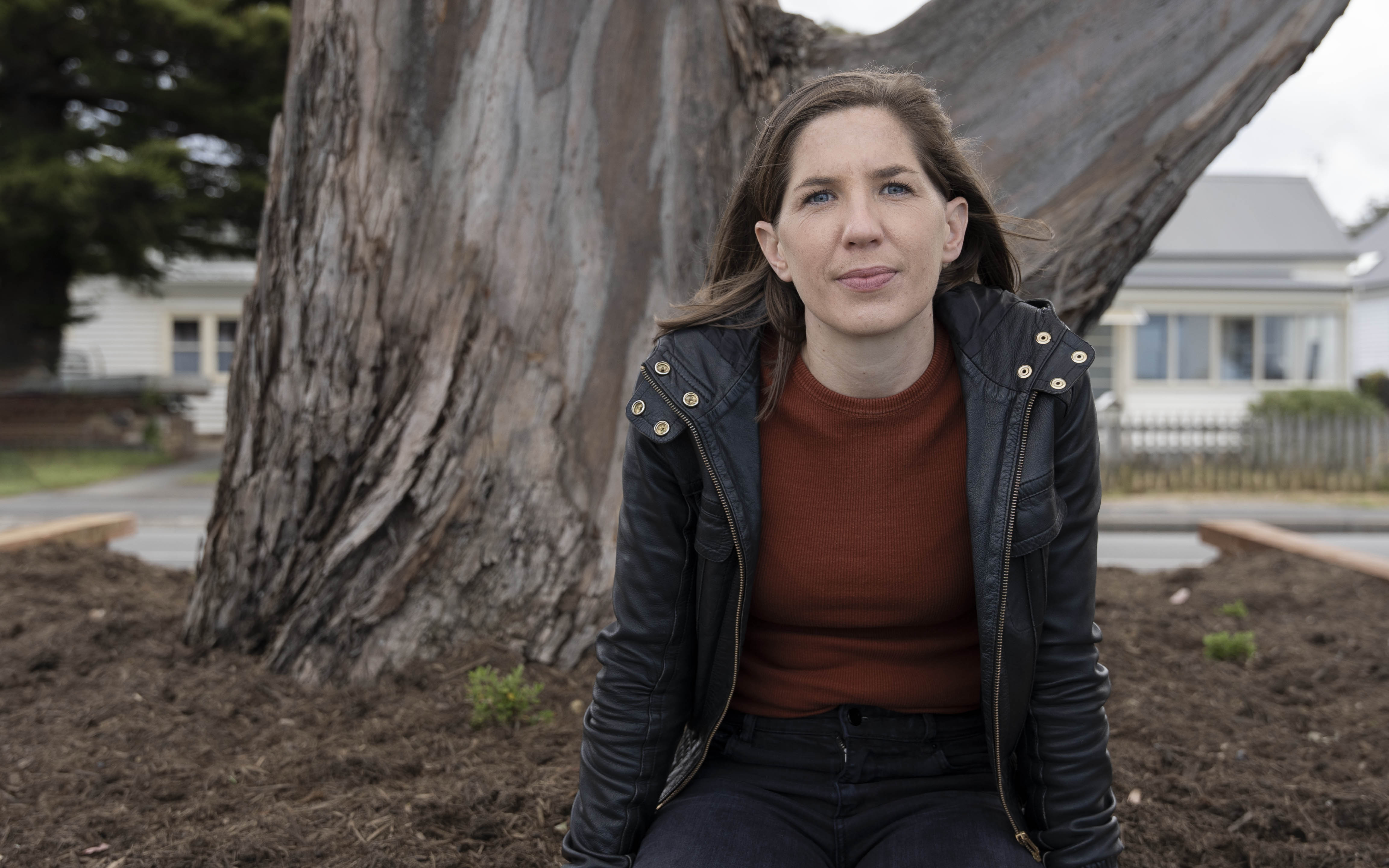 a woman in a black jacket sits in front of a huge tree trunk