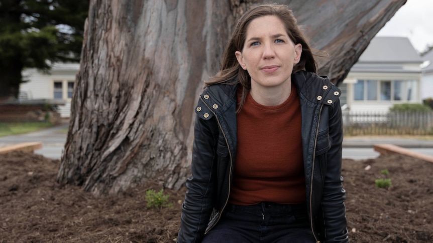 a woman in a black jacket sits in front of a huge tree trunk