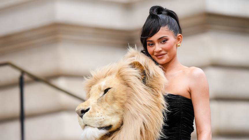 Kylie Jenner - a young woman with black hair in a black dress with a large Lion's head - smiles for the camera