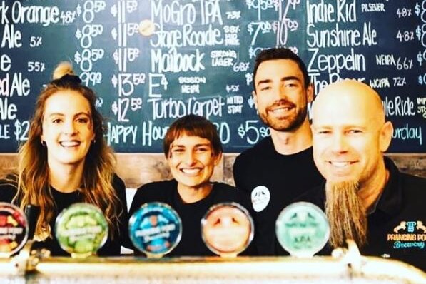 Four brewery workers smile together. They are in front of a 'beers on tap' board and behind beer taps.