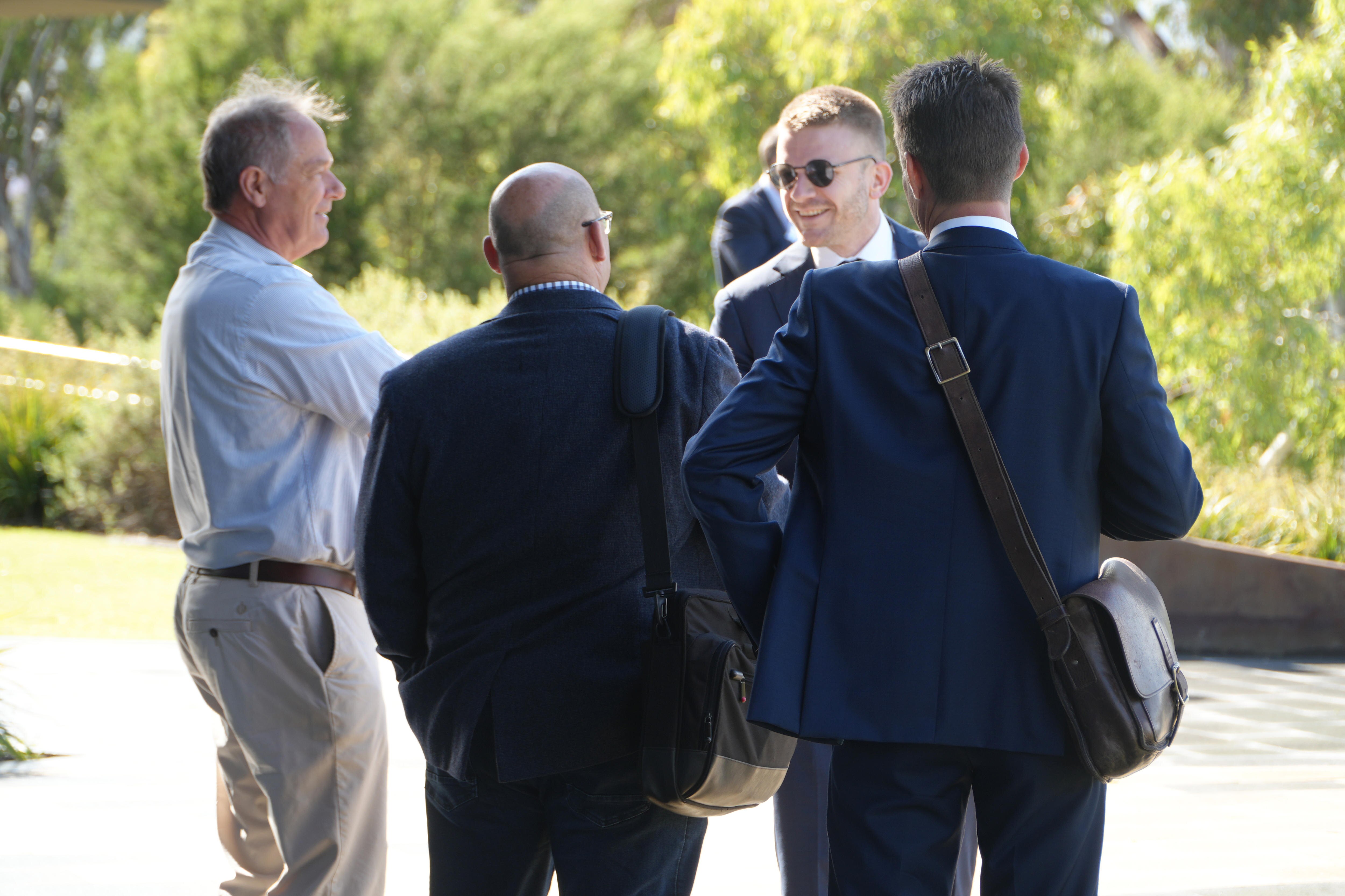 A group of four men wearing suits and business attire stand outside in conversation, one smiling.