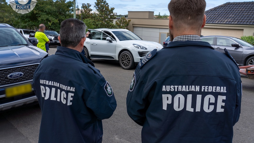 Two police officers in jackets look on as colleagues inspect a white car