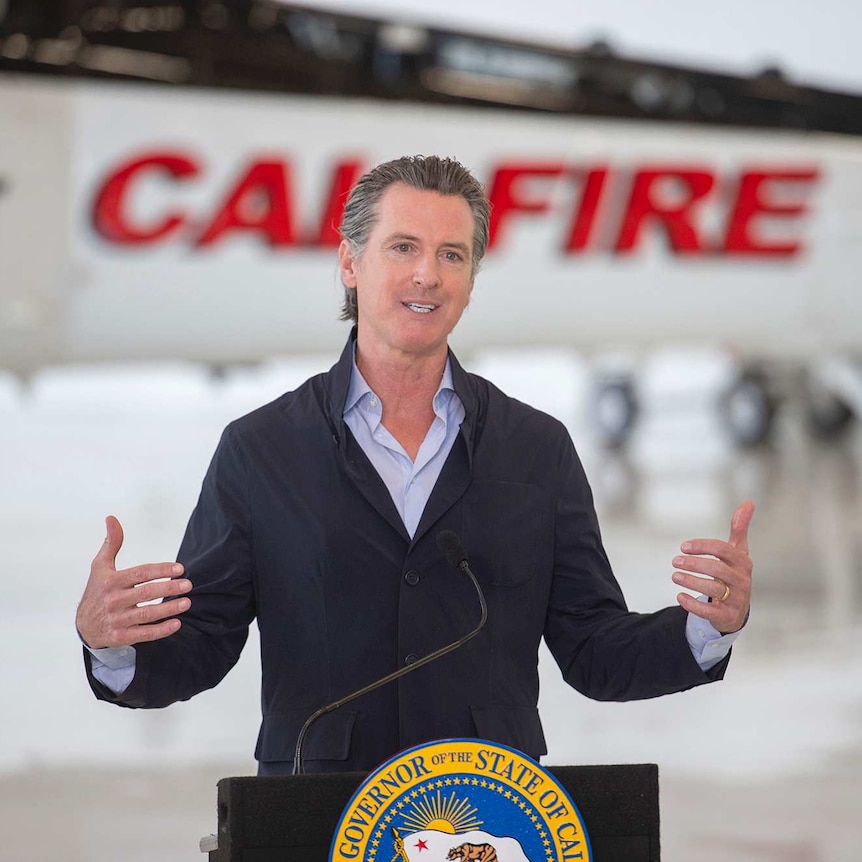 A man gives a speech standing at a podium with an aircraft in the background.