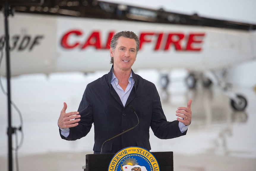 A man gives a speech standing at a podium with an aircraft in the background.