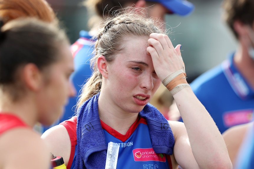 Une joueuse AFLW fatiguée et affectée par la chaleur met sa main sur son front alors qu'elle porte une serviette glacée autour du cou.