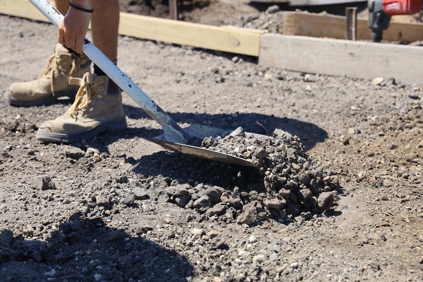 A close up shot of a shovel filled with grey gravel next to sturdy sand- coloured boots