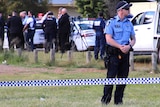 A police officer stands on grass behind police tape, with other officers in the background.