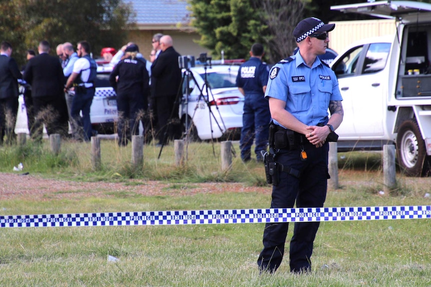 A police officer stands on grass behind police tape, with other officers in the background.