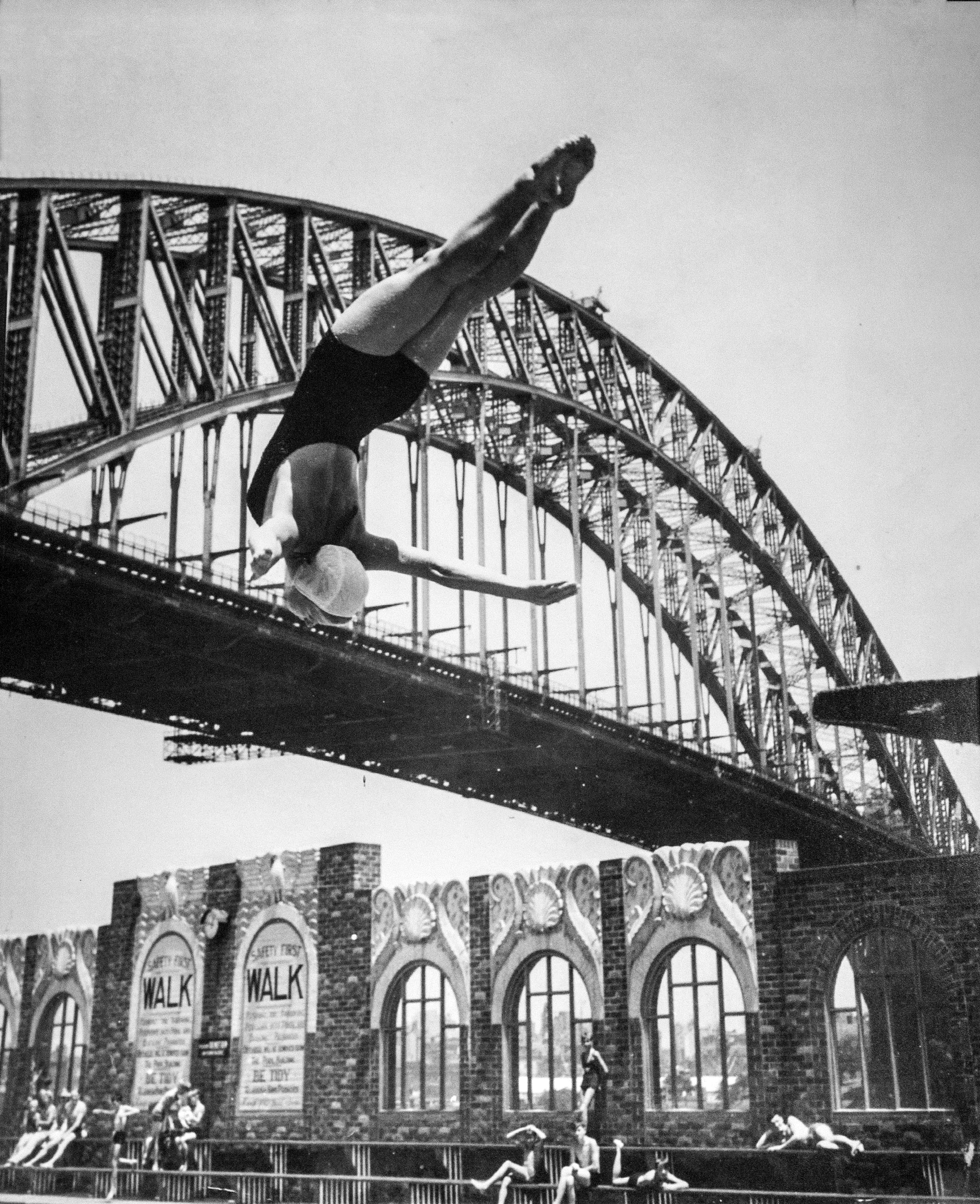 A woman backflips off a diving board in front of the sydney harbour bridge into a pool