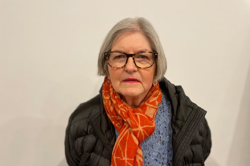 A woman wearing a blue top, black puffer jacket and orange scarf, with short grey hair and glasses
